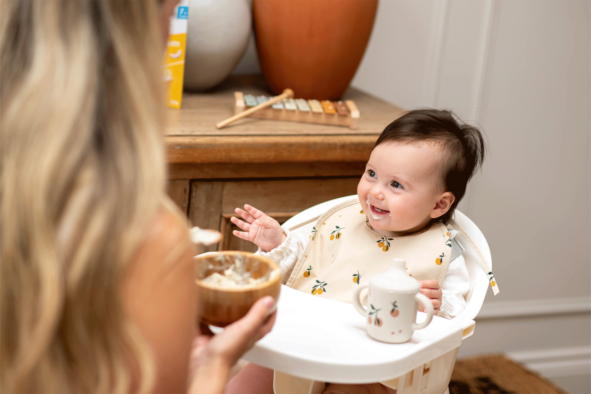 A smiling baby sitting in a high chair wearing a patterned bib, looking up at a mother holding a bowl of food.