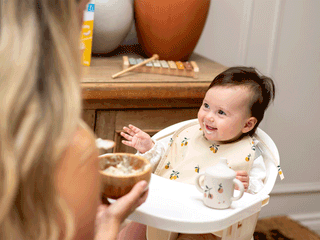 A smiling baby sitting in a high chair wearing a patterned bib, looking up at a mother holding a bowl of food.