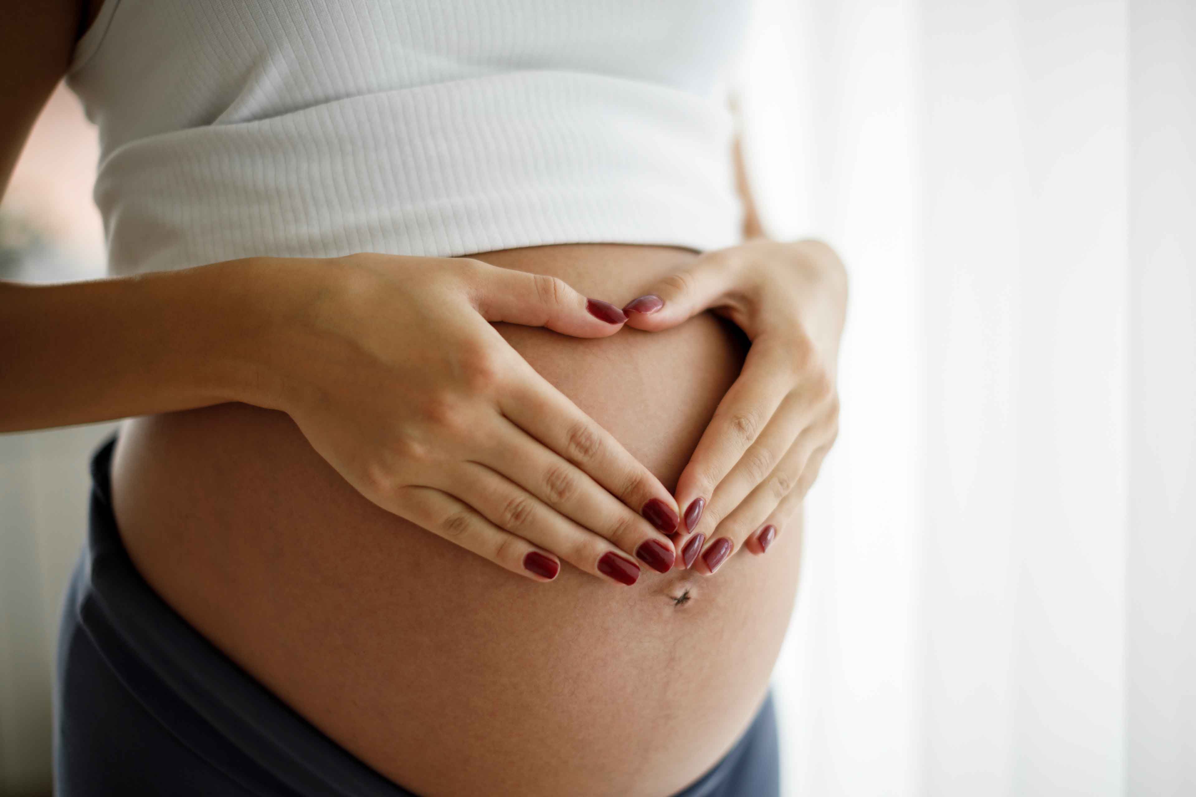 Schwangere Frau legt Hände mit roten Fingernägeln in Herzform auf ihren nackten Bauch
