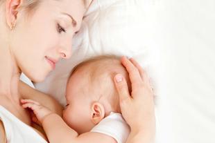 Mutter liegt entspannt im Bett und stillt ihr Neugeborenes, hält dabei schützend den Kopf des Babys