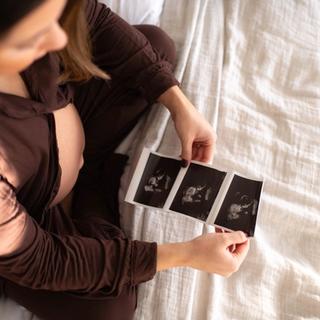 Pregnant woman sitting on a bed, holding and looking at ultrasound images of her baby.