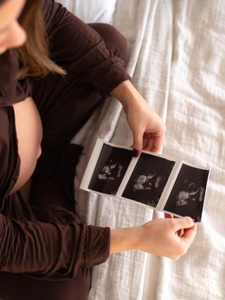 Pregnant woman sitting on a bed, holding and looking at ultrasound images of her baby.