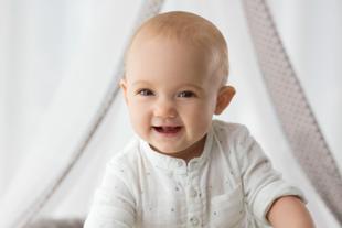 Lachendes Baby in heller Kleidung sitzt aufrecht und wirkt zufrieden und gesund.
