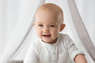Lachendes Baby in heller Kleidung sitzt aufrecht und wirkt zufrieden und gesund.