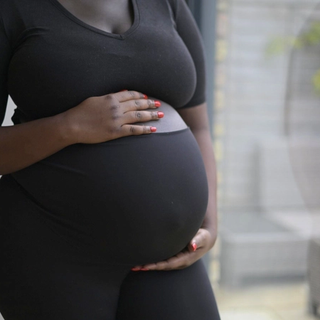 A pregnant woman wearing a fitted black outfit cradles her belly with both hands. Her nails are painted a vibrant red, contrasting with her dark clothing. The background features a softly lit outdoor patio with wicker furniture and plants, slightly blurred.