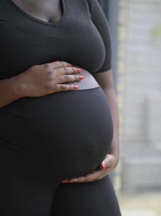 A pregnant woman wearing a fitted black outfit cradles her belly with both hands. Her nails are painted a vibrant red, contrasting with her dark clothing. The background features a softly lit outdoor patio with wicker furniture and plants, slightly blurred.