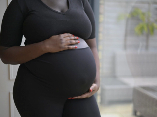 A pregnant woman wearing a fitted black outfit cradles her belly with both hands. Her nails are painted a vibrant red, contrasting with her dark clothing. The background features a softly lit outdoor patio with wicker furniture and plants, slightly blurred.