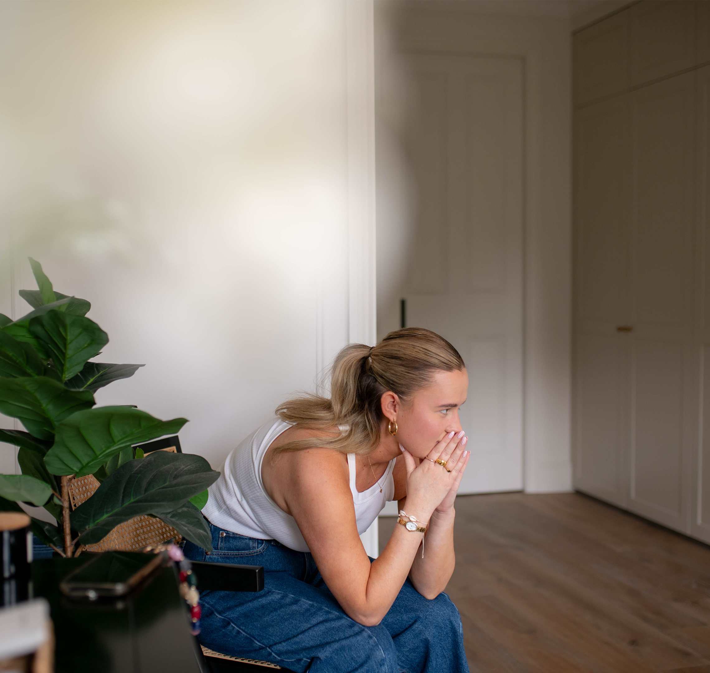 Pregnant woman sitting on a chair at home, leaning forward with hands to her mouth, looking worried and unwell.
