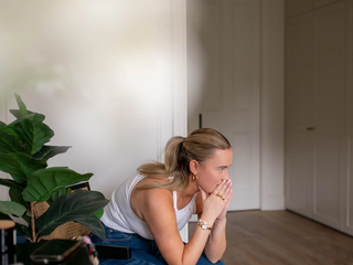 Pregnant woman sitting on a chair at home, leaning forward with hands to her mouth, looking worried and unwell.
