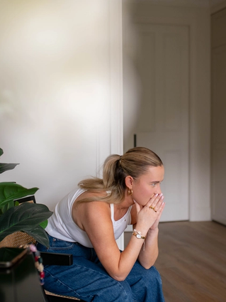 Pregnant woman sitting on a chair at home, leaning forward with hands to her mouth, looking worried and unwell.
