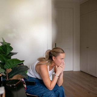 Pregnant woman sitting on a chair at home, leaning forward with hands to her mouth, looking worried and unwell.