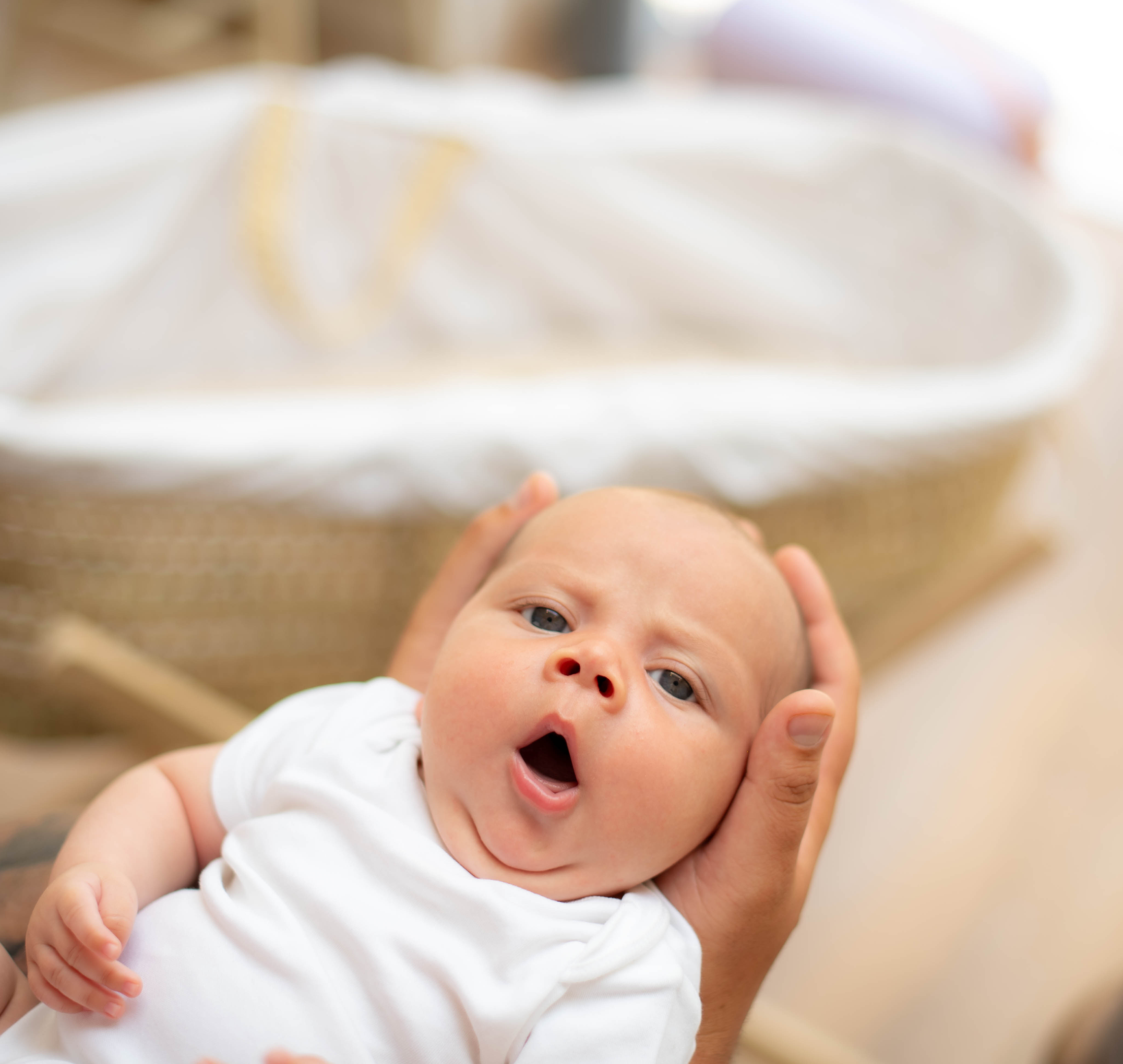 A young baby in a white babygrow being gently held, looking up with its mouth open as if yawning, with a soft-focus Moses basket in the background.