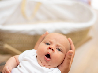 A young baby in a white babygrow being gently held, looking up with its mouth open as if yawning, with a soft-focus Moses basket in the background.