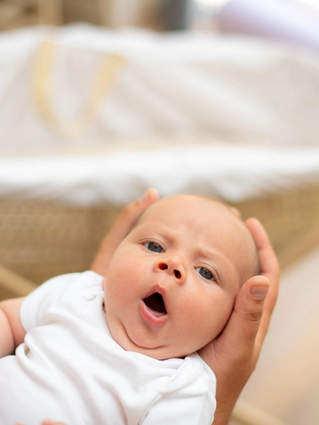 A young baby in a white babygrow being gently held, looking up with its mouth open as if yawning, with a soft-focus Moses basket in the background.