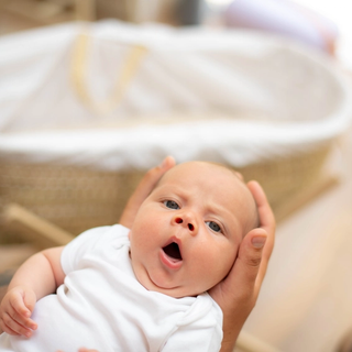 A young baby in a white babygrow being gently held, looking up with its mouth open as if yawning, with a soft-focus Moses basket in the background.