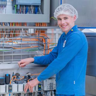 Auszubildender Mechatroniker arbeitet lächelnd an einer Schalttafel mit Kabeln und Steuerungstechnik in der Produktion.