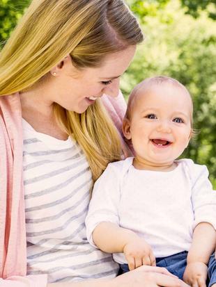 Frau mit langem Haar in gestreiftem Shirt und rosa Jacke hält Baby in weißem Oberteil auf dem Schoß im Freien