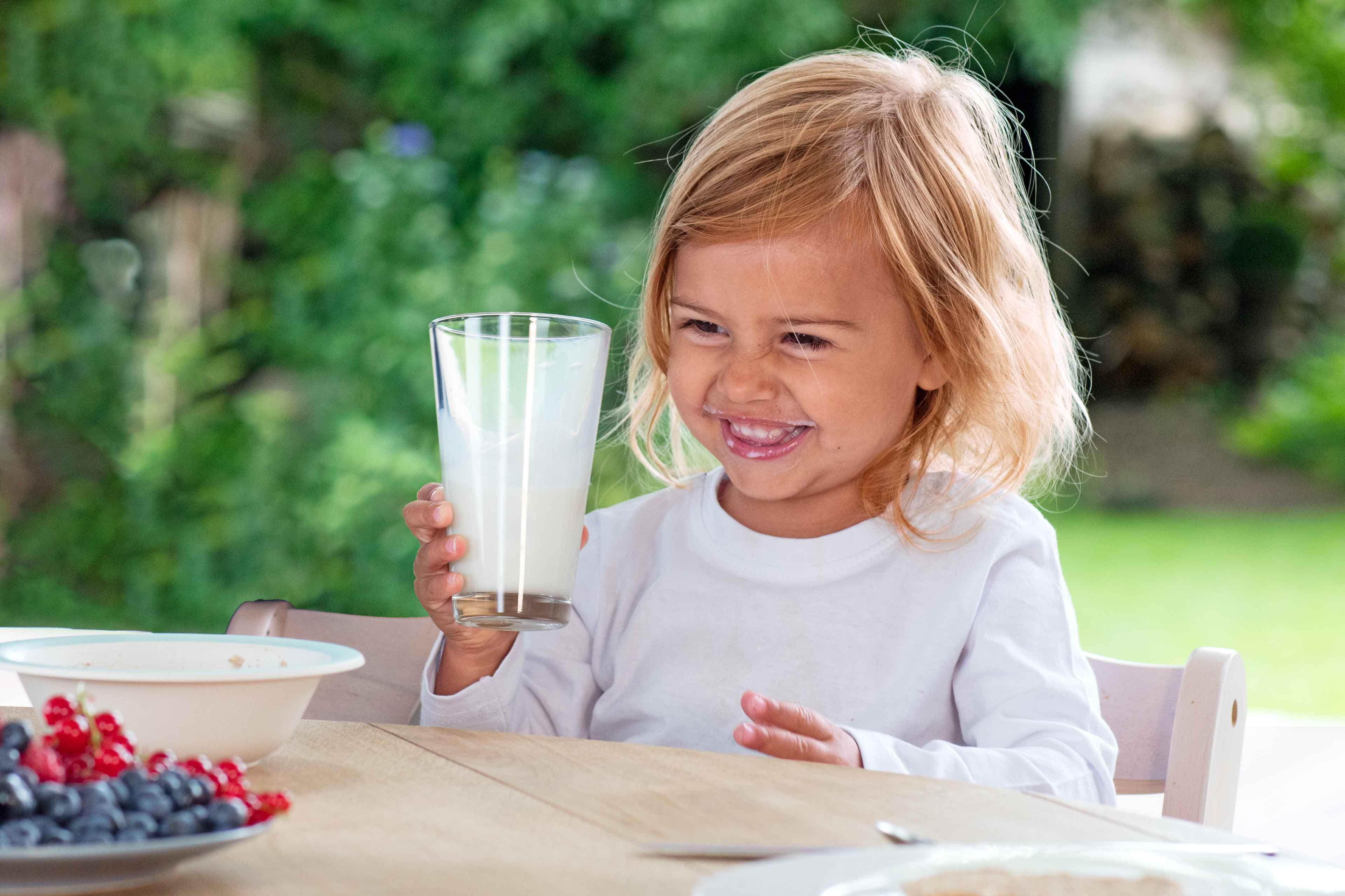 Ein fröhliches Kind hält ein Glas mit weißer Flüssigkeit, vermutlich Milch, in der Hand.