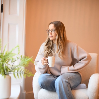 Woman sitting in a cozy chair by the window, holding a mug and gazing outside thoughtfully.