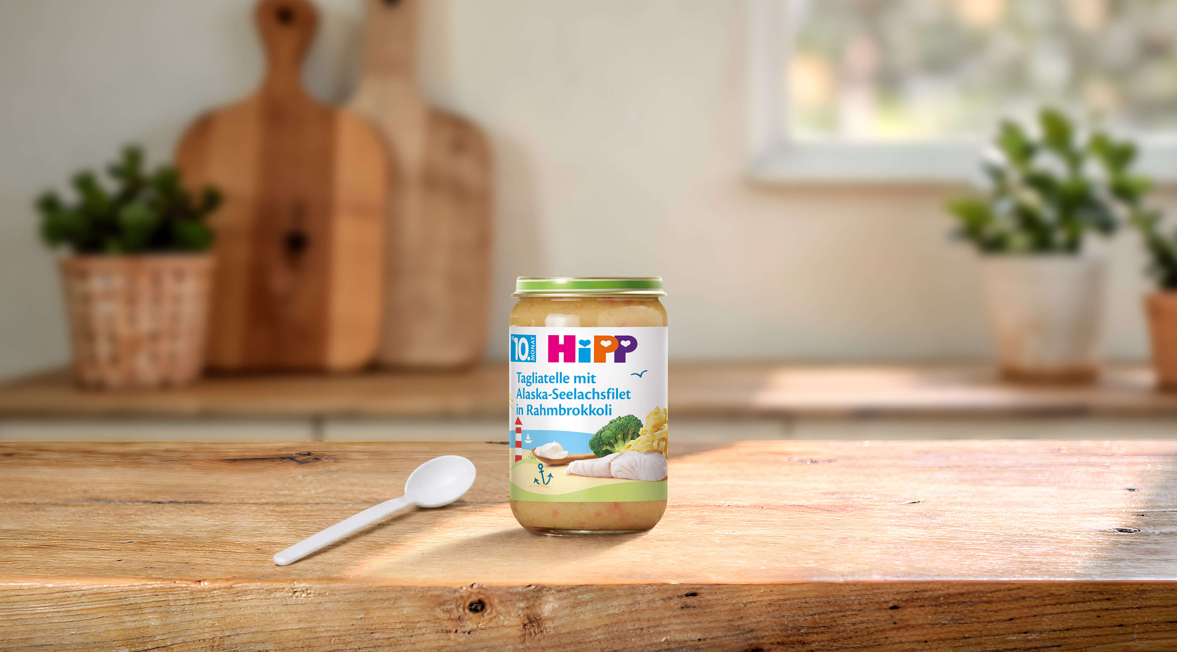 HiPP Bio Babyglas Tagliatelle mit Alaska-Seelachsfilet in Rahmbrokkoli ab dem 10. Monat auf Holztisch mit Löffel in heller Küche