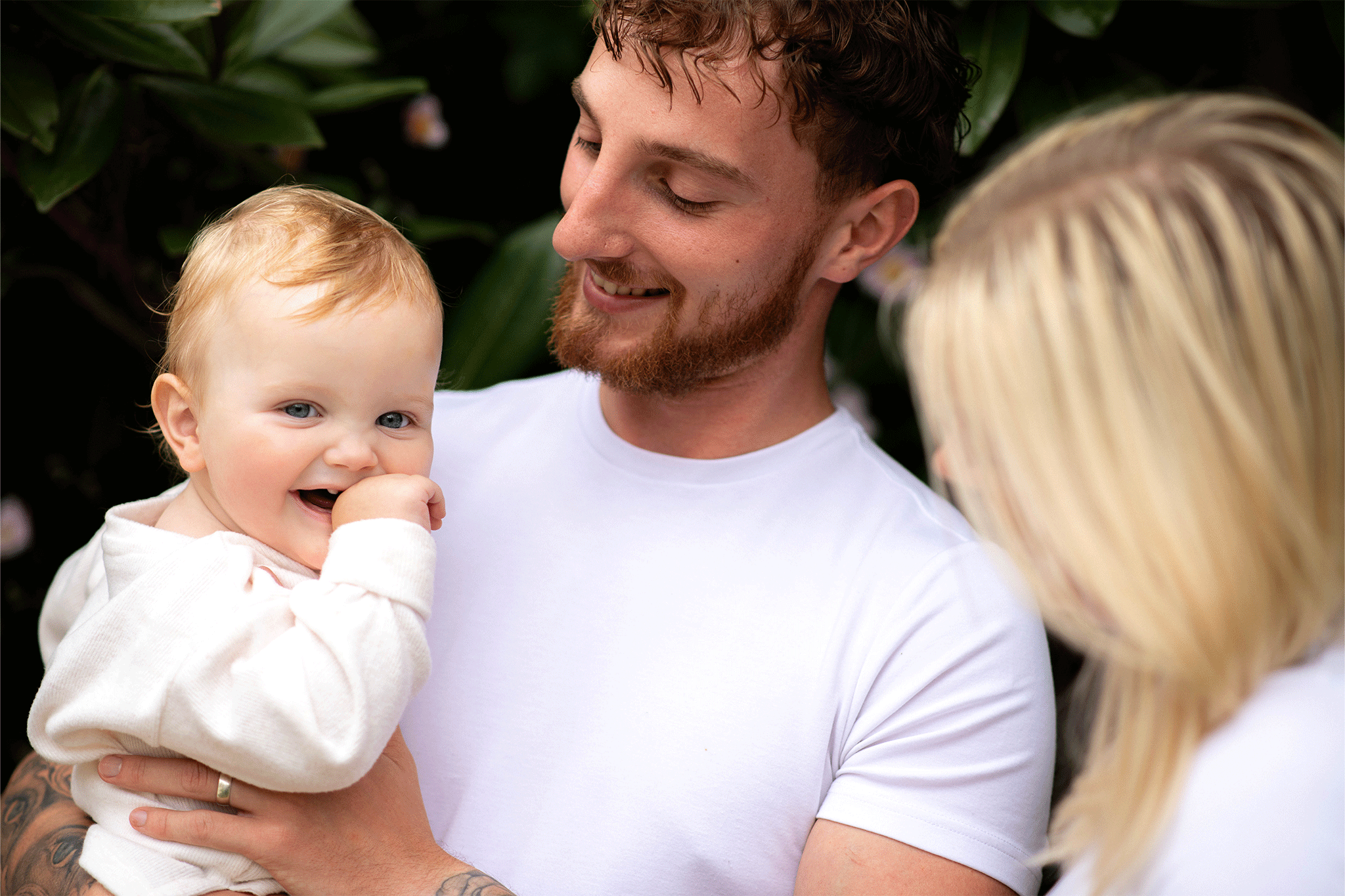 A young family shares a candid moment outdoors against a background of lush green foliage. On the left, a man with light brown wavy hair and a trimmed beard, wearing a white t-shirt, holds a smiling baby with fair skin and short light reddish-blonde hair. The baby, dressed in a light-colored top, looks toward the camera with a hand near their mouth. To the right, seen from the side and slightly behind, a woman with straight blonde hair looks toward the man and baby.