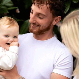 A young family shares a candid moment outdoors against a background of lush green foliage. On the left, a man with light brown wavy hair and a trimmed beard, wearing a white t-shirt, holds a smiling baby with fair skin and short light reddish-blonde hair. The baby, dressed in a light-colored top, looks toward the camera with a hand near their mouth. To the right, seen from the side and slightly behind, a woman with straight blonde hair looks toward the man and baby.