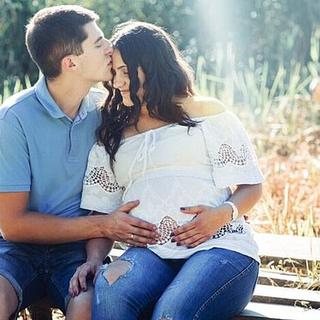 Expectant couple sitting on a bench outdoors, with the man kissing the woman's forehead and both hands resting on her pregnant belly.