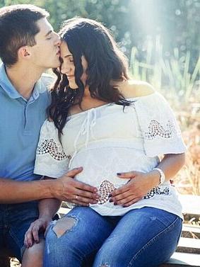 Expectant couple sitting on a bench outdoors, with the man kissing the woman's forehead and both hands resting on her pregnant belly.