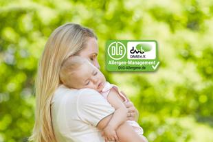 Mutter hält ihr schlafendes Baby im Grünen im Arm, daneben ein grünes DLG-/DAAB-Allergen-Management-Siegel.