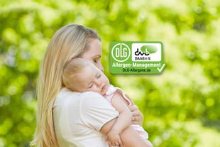 Mutter hält ihr schlafendes Baby im Grünen im Arm, daneben ein grünes DLG-/DAAB-Allergen-Management-Siegel.