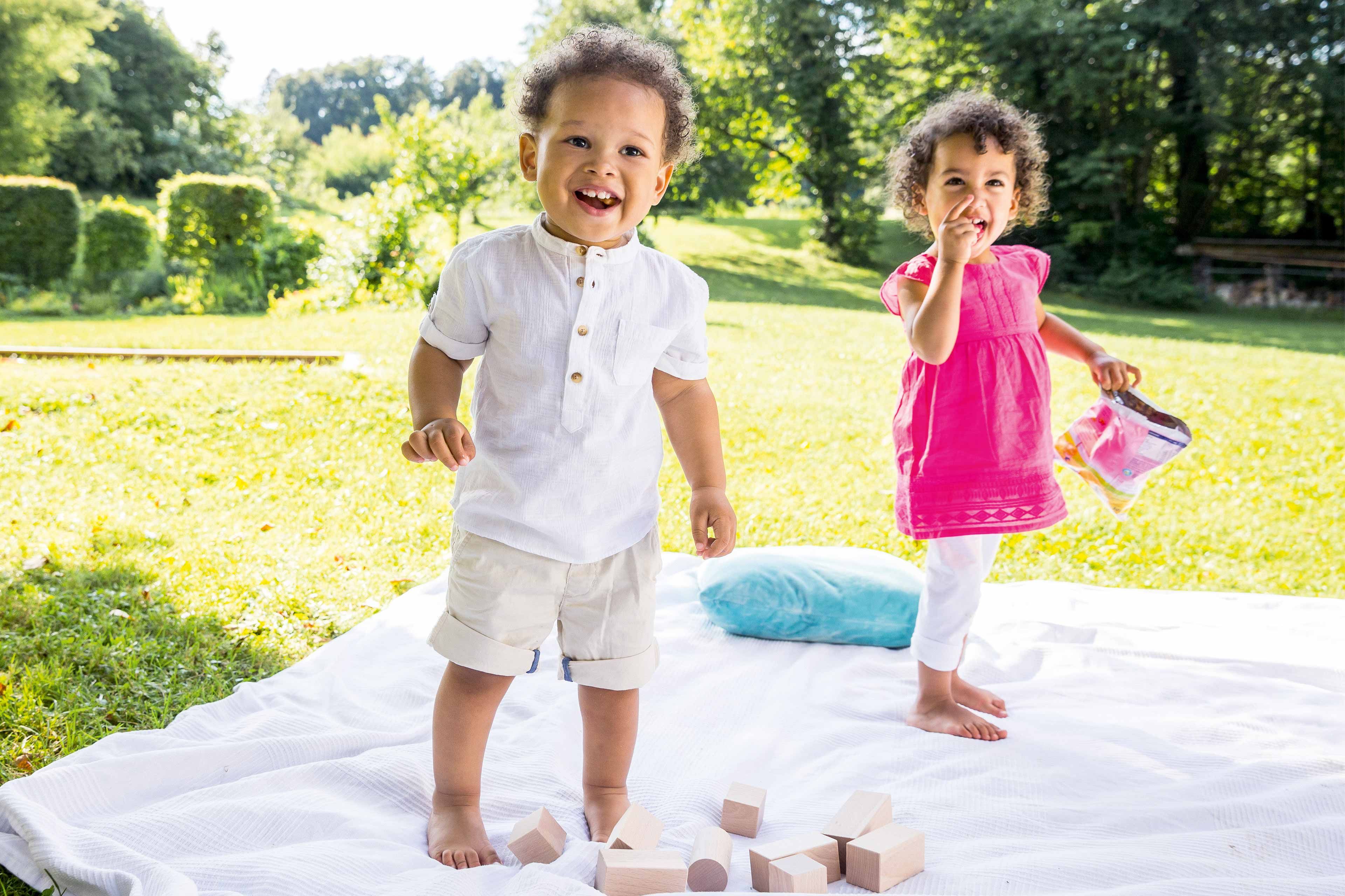 Zwei kleine Kinder spielen im Freien auf einer Decke, umgeben von Holzspielzeug und Grünzeug.