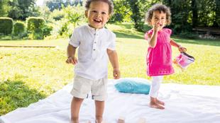 Zwei kleine Kinder spielen im Freien auf einer Decke, umgeben von Holzspielzeug und Grünzeug.