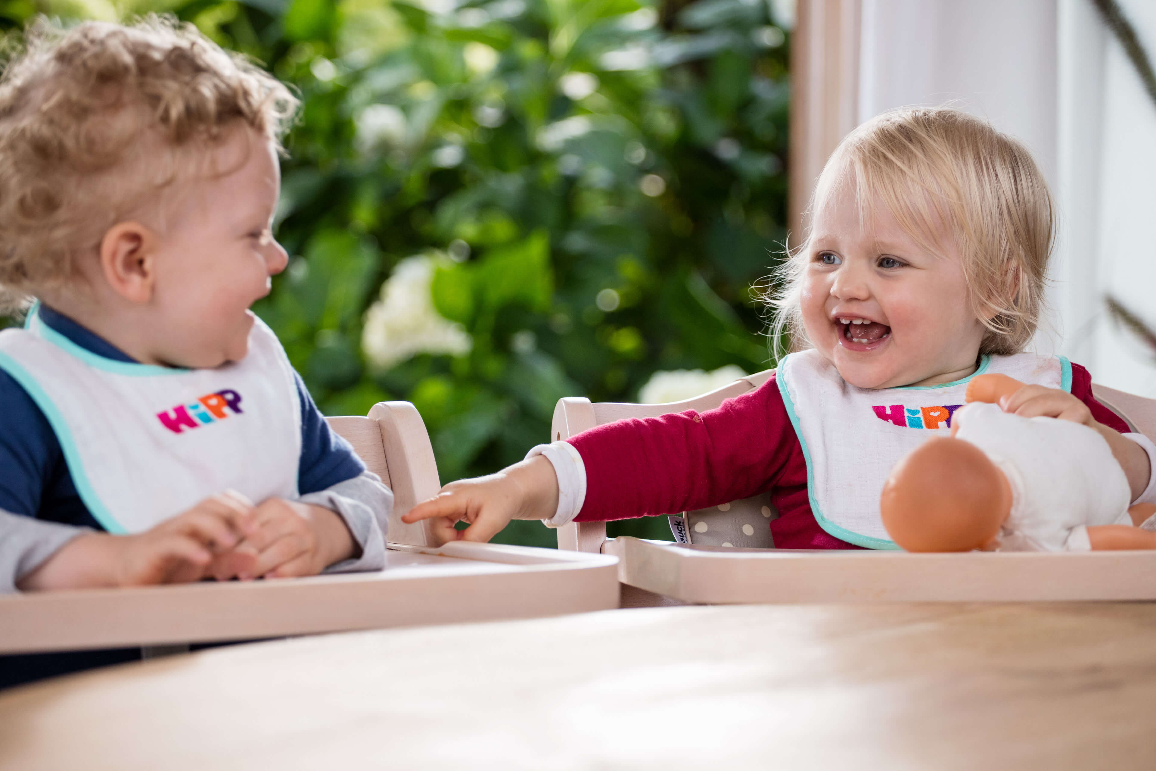 Zwei Kleinkinder, eines mit lockigem und das andere mit blondem Haar, sitzen beide in Hochstühlen, lachen und spielen mit einer Puppe.