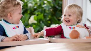 Zwei Kleinkinder, eines mit lockigem und das andere mit blondem Haar, sitzen beide in Hochstühlen, lachen und spielen mit einer Puppe.