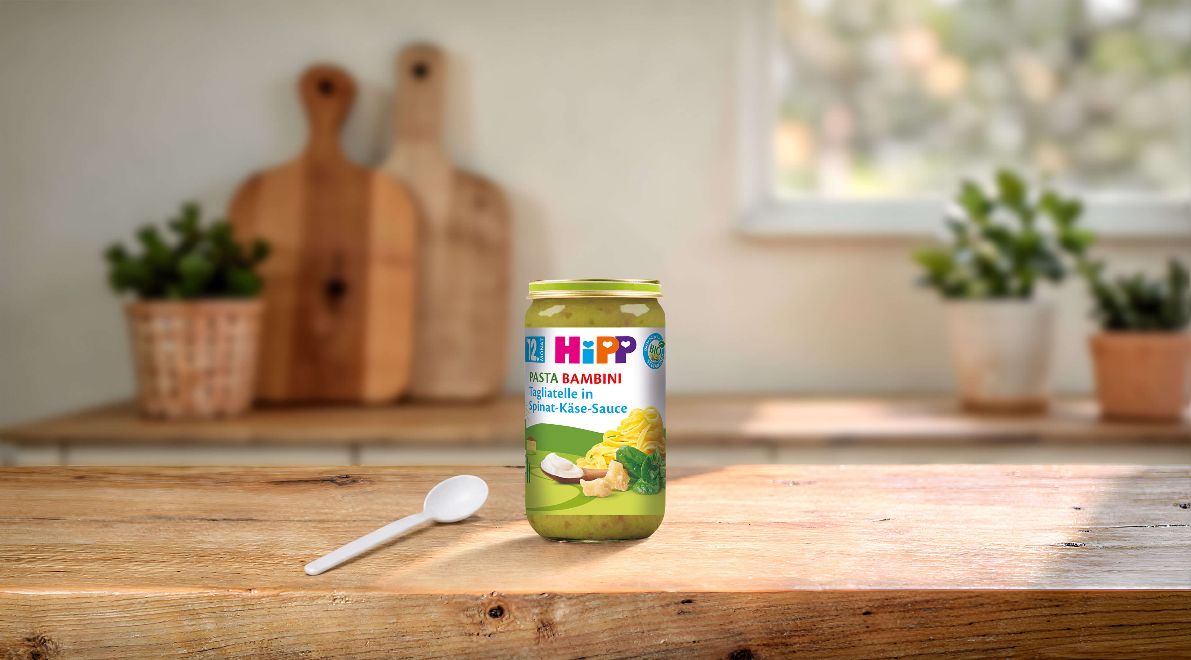 HiPP Pasta Bambini Glas Tagliatelle in Spinat-Käse-Sauce ab 12 Monaten auf Holztisch mit Löffel in heller Küche