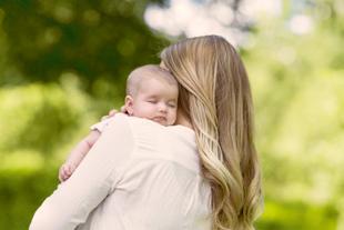Eine Frau mit langen blonden Haaren, die ein schlafendes Baby vor einem grünen Hintergrund hält.