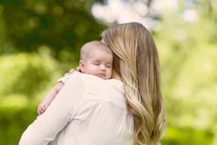 Eine Frau mit langen blonden Haaren, die ein schlafendes Baby vor einem grünen Hintergrund hält.