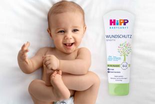 Ein Baby mit freudigem Gesichtsausdruck, das auf einer weißen Fläche liegt, neben einer Tube "WUNDSCHUTZ SENSITIV"-Creme, die mit "Mit Bio-Mandelblend", "21% Panthenol" und "Parfumfrei" gekennzeichnet ist.