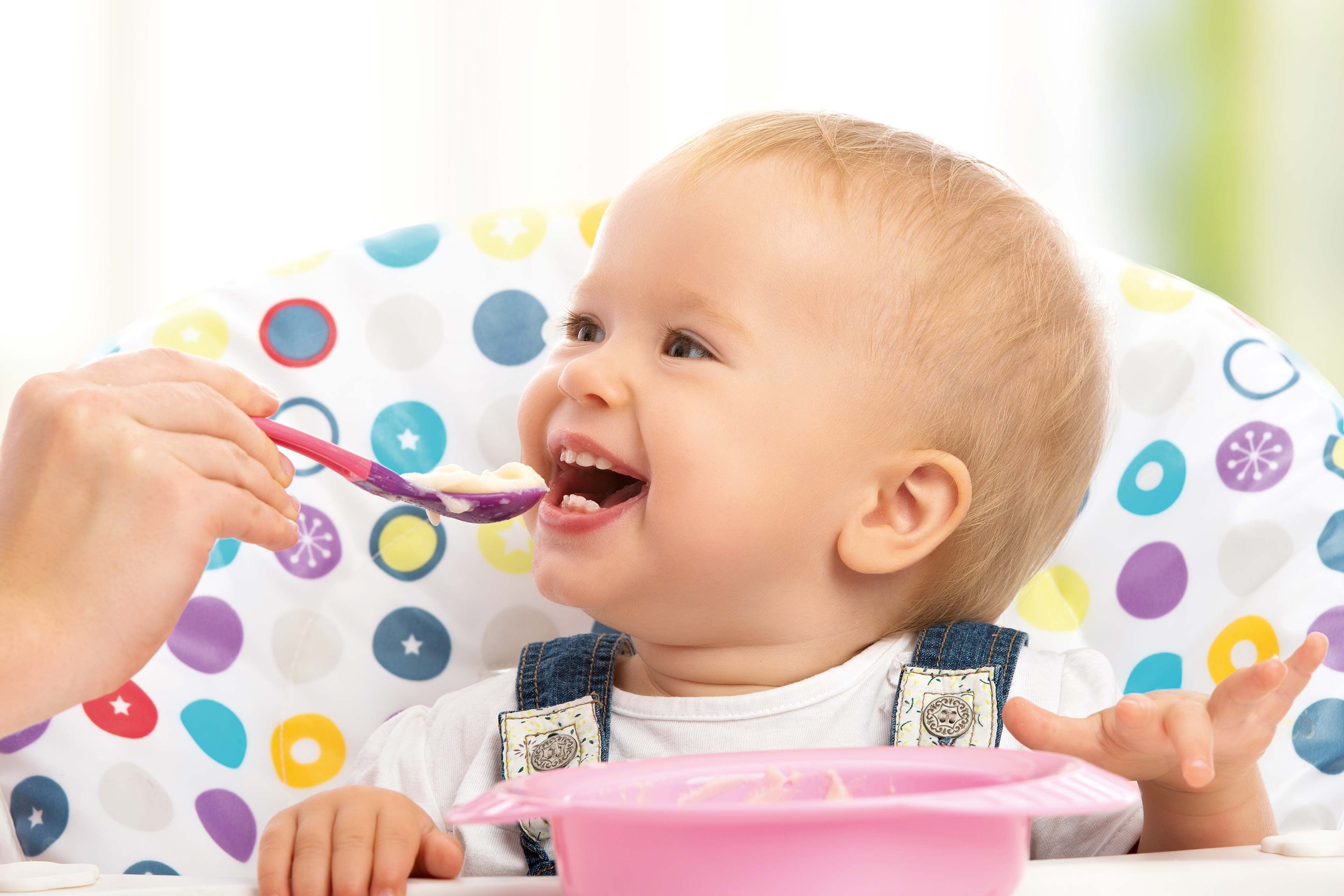 Ein Baby, das von einem Erwachsenen mit einem Löffel gefüttert wird, sitzt auf einem Stuhl mit einem bunten Tupfenmuster.