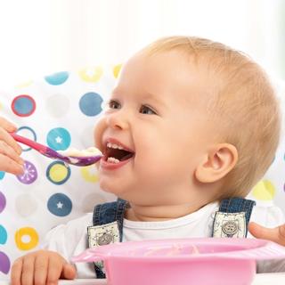 Ein Baby, das von einem Erwachsenen mit einem Löffel gefüttert wird, sitzt auf einem Stuhl mit einem bunten Tupfenmuster.