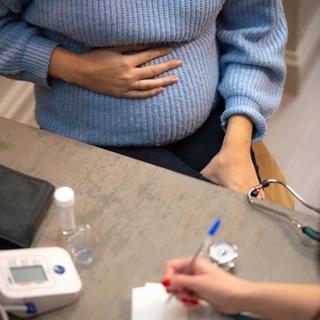 Close-up of a pregnant woman holding her belly during a prenatal check-up, with medical equipment and a healthcare professional writing notes nearby.
