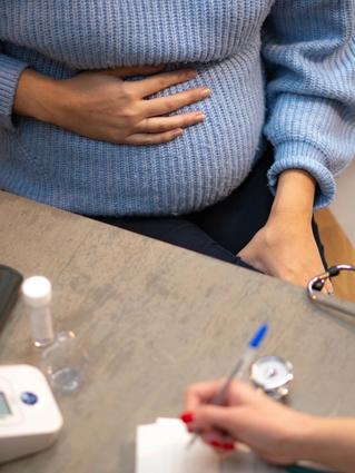 Close-up of a pregnant woman holding her belly during a prenatal check-up, with medical equipment and a healthcare professional writing notes nearby.
