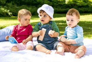 Drei Babys sitzen im Freien auf einer Decke, jedes hält ein Spielzeug.