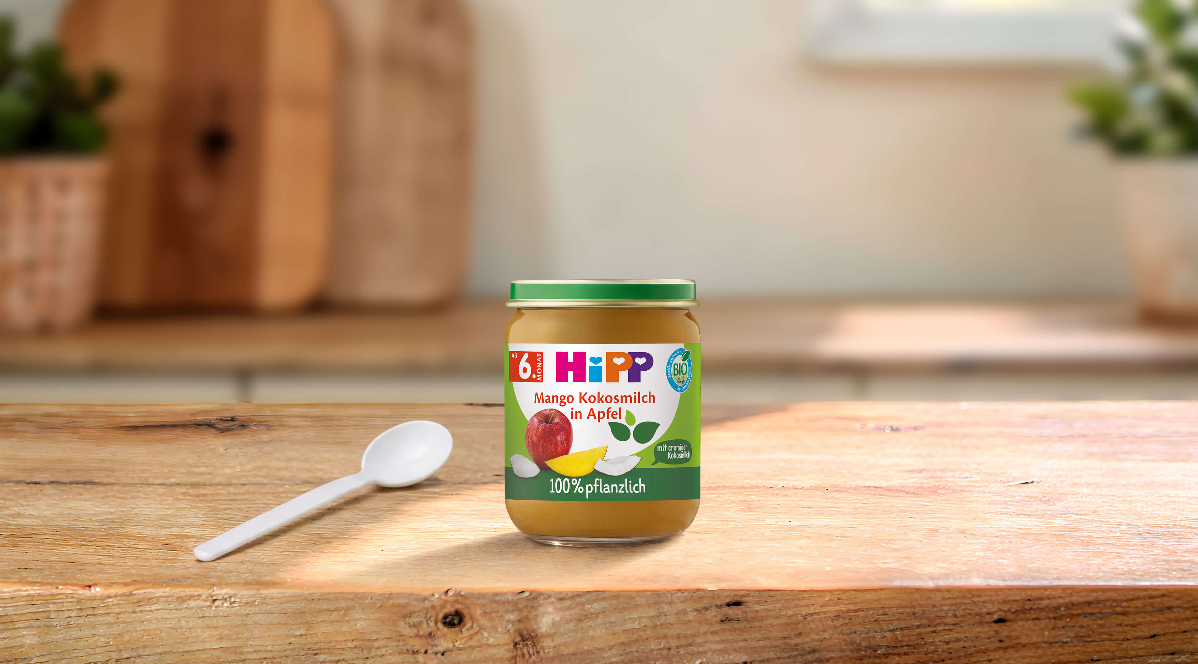 HiPP Bio Babygläschen Mango Kokosmilch in Apfel, 100 % pflanzlich, auf Holztisch mit Löffel in heller Küchenumgebung