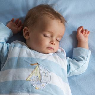 Ein scheinbar schlafendes Baby in einem blau-weiß gestreiften Outfit mit Segelbootmotiv ruht auf einer blauen Fläche.