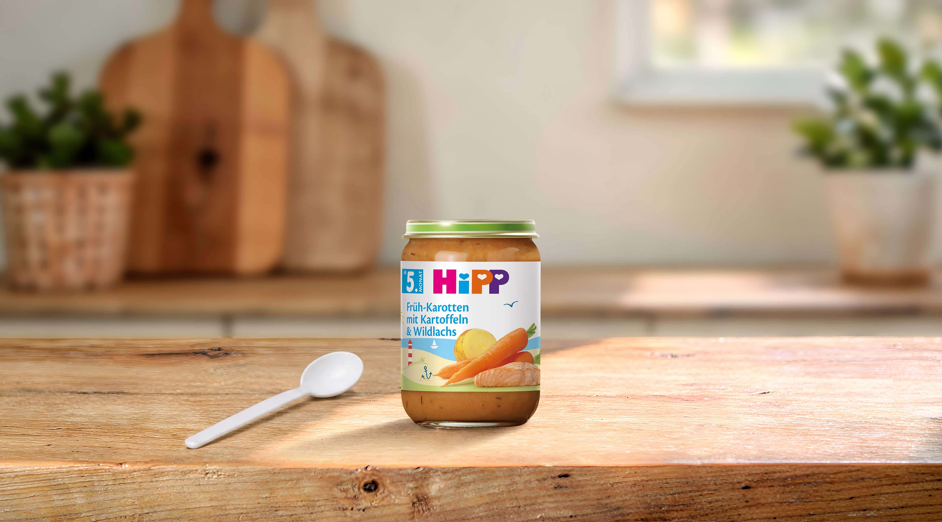 HiPP Früh-Karotten mit Kartoffeln & Wildlachs Bio-Babygläschen auf Holztisch mit Löffel in heller Küche
