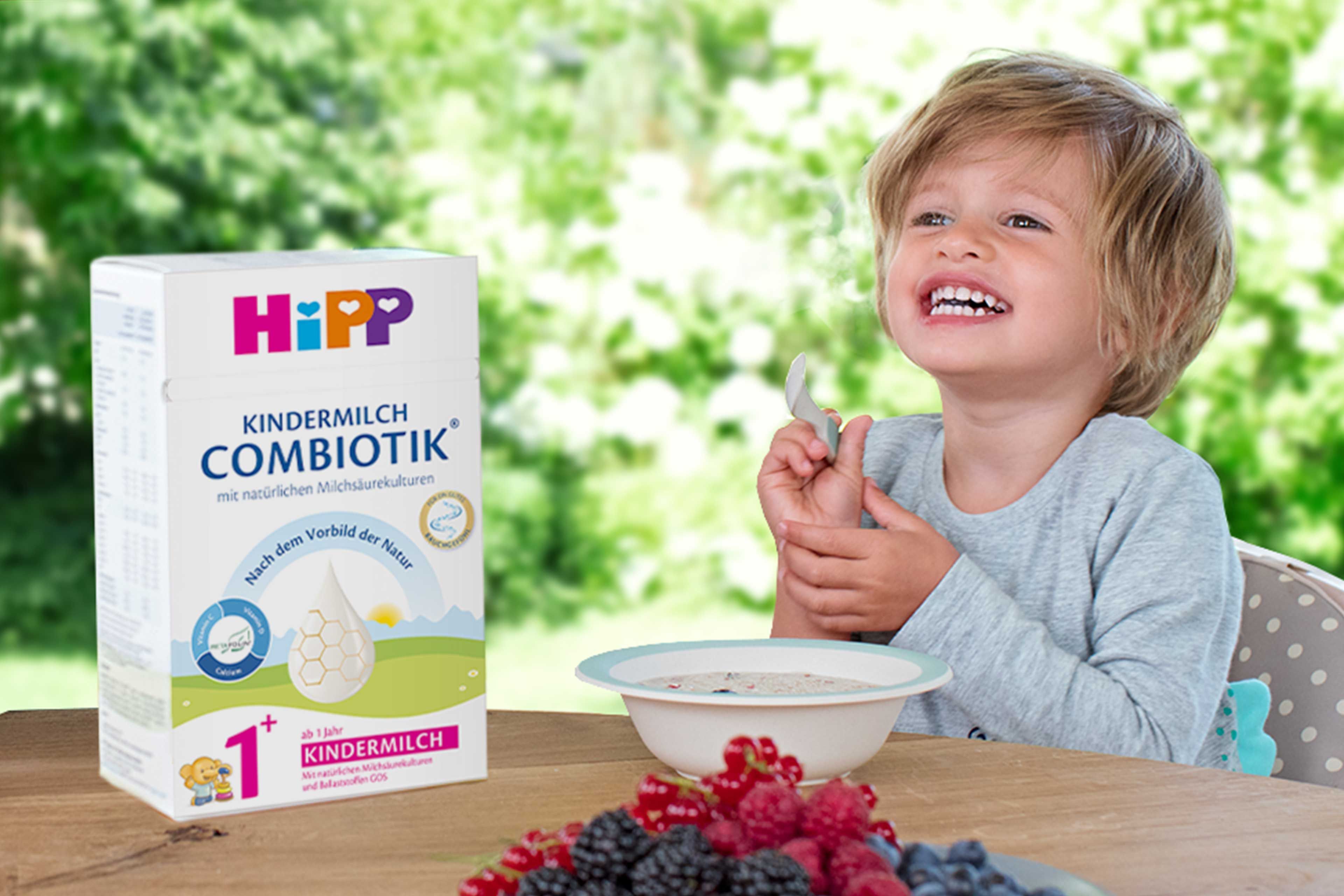 Kind sitzt am Tisch mit einer Schale und Löffel, daneben steht eine Packung HiPP Kindermilch Combiotik, im Vordergrund liegen Beeren.