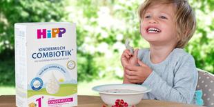 Kind sitzt am Tisch mit einer Schale und Löffel, daneben steht eine Packung HiPP Kindermilch Combiotik, im Vordergrund liegen Beeren.