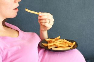Eine Person in einem rosa Hemd, die einen Teller mit Pommes frites in der Hand hält und einen sichtbaren Schwangerschaftsbauch hat.