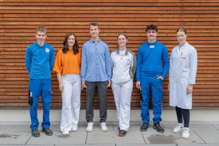 Sechs junge Erwachsene stehen nebeneinander vor einer Holzfassade, zwei tragen blaue Arbeitskleidung, zwei weiße Arbeitsjacken, eine Person einen orangen Pullover und eine Person ein blaues Hemd
